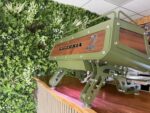Green coffee machine on wooden counter with greenery backdrop.