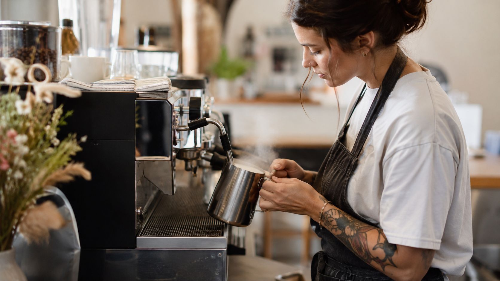 A barista with tattoos steaming plant-based milk using an espresso machine in a modern coffee shop.