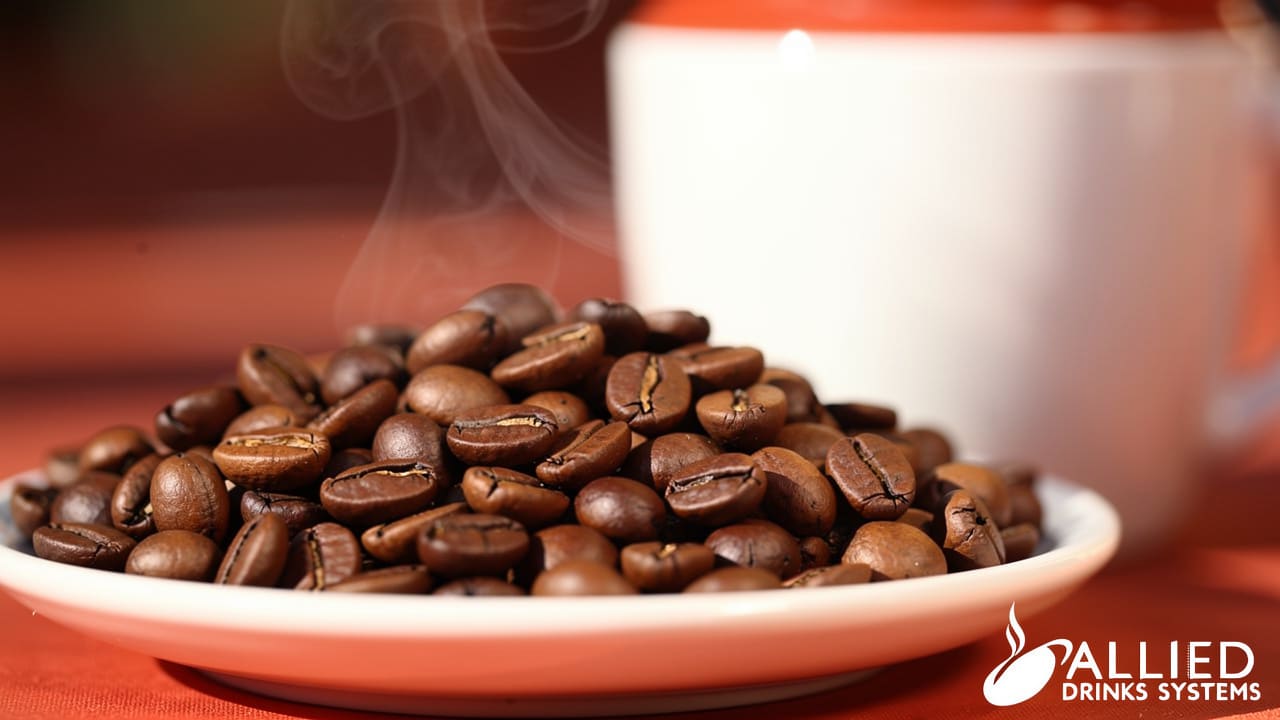 Dark roasted coffee beans piled on a white plate with a steaming cup blurred in the background against an orange surface.
