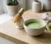 Matcha set on a pale wooden counter: ceramic bowl of frothy matcha, bamboo whisk on a stand, sieve and scoop